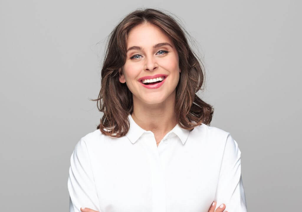 Woman with full arch implants smiling wearing a white shirt against a grey background